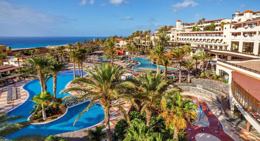 High-angle view of a hotel resort showing winding blue swimming pools with footbridges, palm trees, sun loungers, and white buildings with an ocean horizon