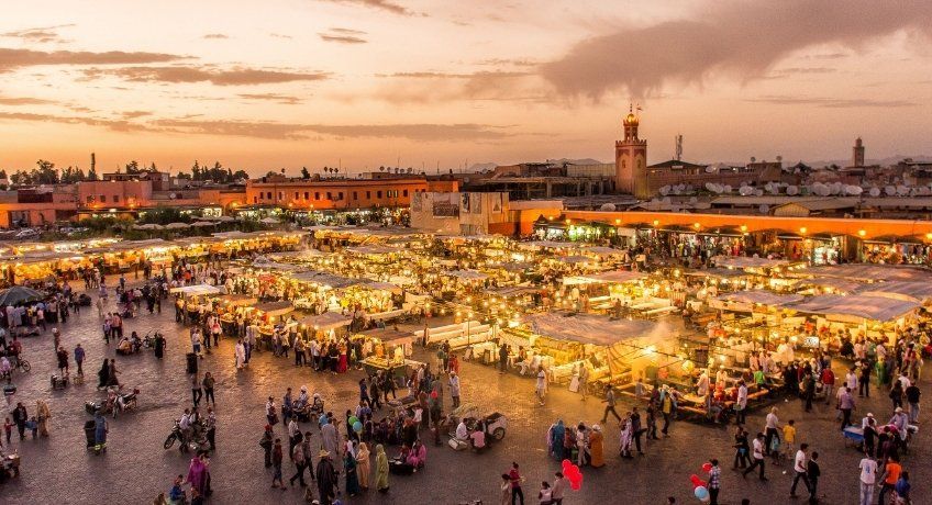 Aerial view of a night market plaza: vendor stalls, crowds under warm lights, dusky orange sky, minarets nearby.