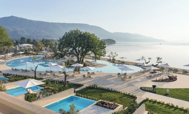Resort terrace overlooking a calm bay and mountains, with multiple swimming pools, sun loungers, umbrellas, landscaped gardens, and a sandy beach along the shoreline.