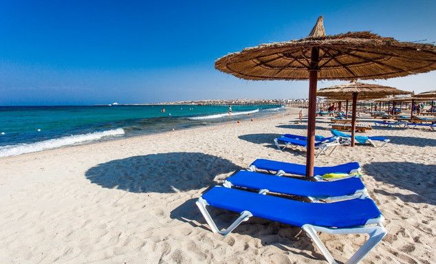 Blue-white striped sunbed and straw umbrella on a sandy beach, calm ocean, distant coastline, clear sky.