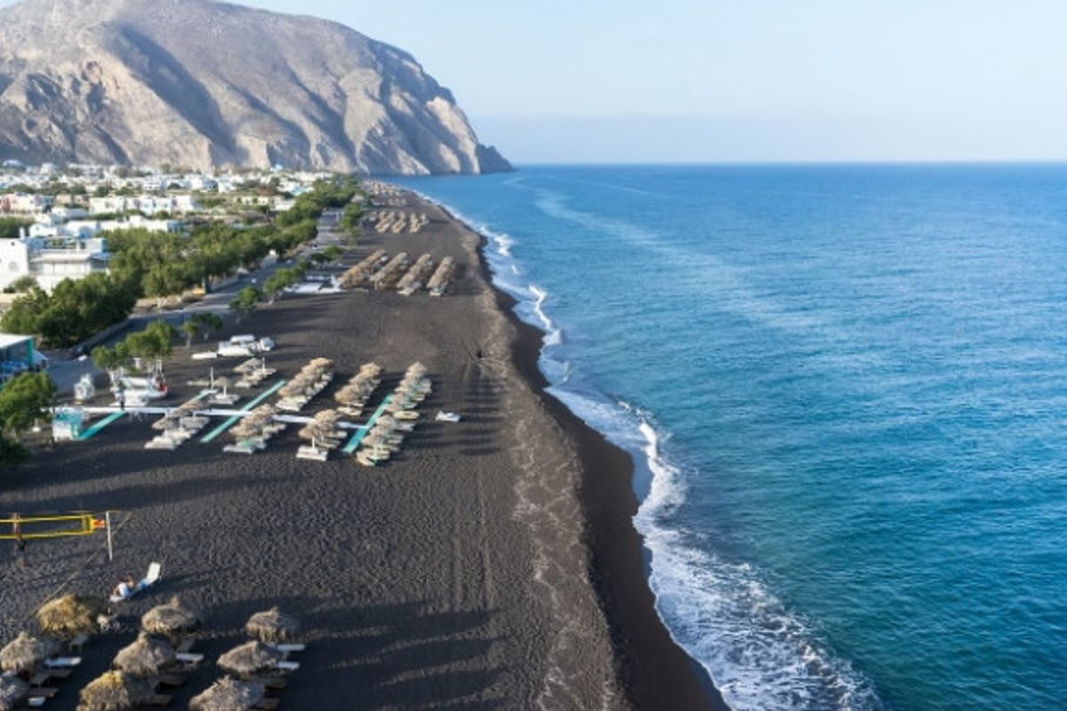 The volcanic black sands of Perissa Beach in Santorini lined with sunloungers and backed by whitewashed houses