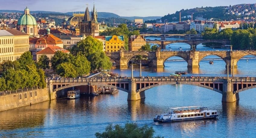 Panoramic riverside city: stone bridge, calm blue water, boats cruising, historic spires and domes, clear sky.