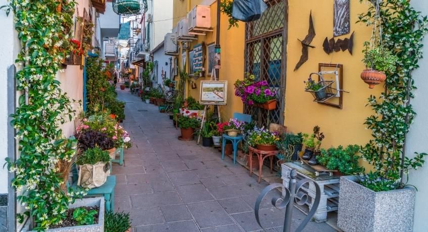 Narrow European alley: potted flowers, teal chairs, hanging lanterns, yellow walls, decorative wall art.