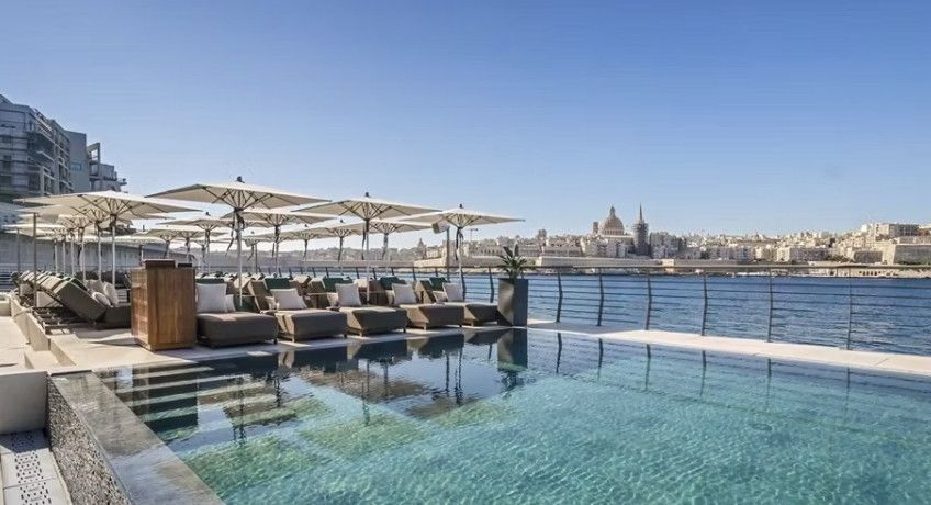 Waterfront infinity pool with sun loungers and white parasols, overlooking the harbour and a domed city skyline.