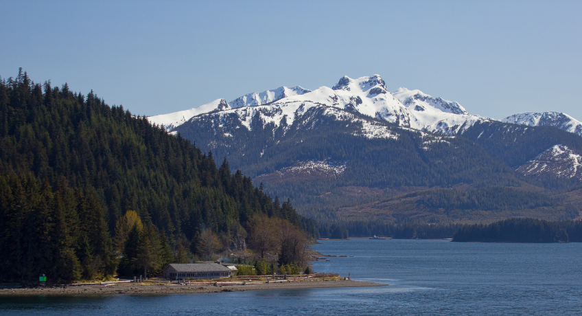 A calm lake shoreline with a small dock and buildings, backed by dark evergreen forest and snow-capped mountains under a clear blue sky.