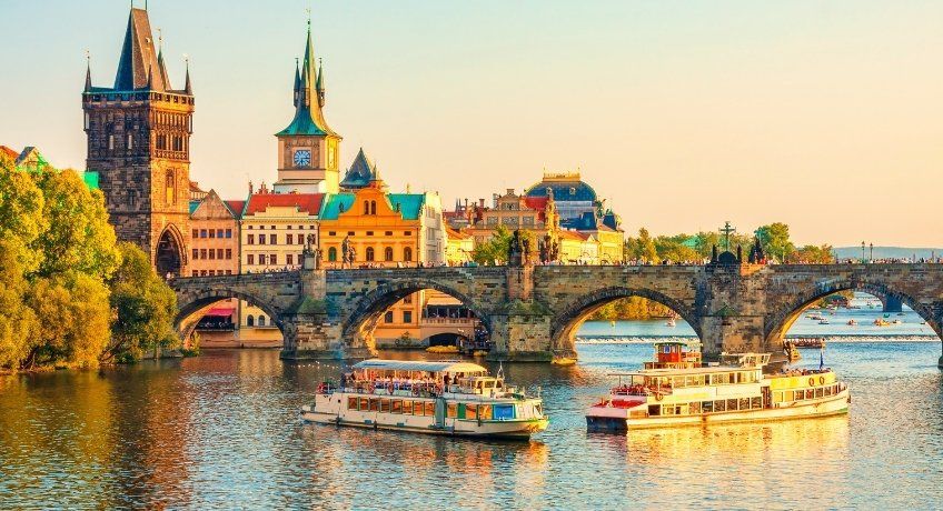 Historic riverside at sunset: cathedral with green spire, stone bridge, sightseeing boats cruising the water.