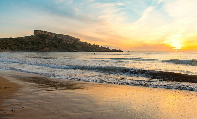 Sunset over a sandy beach with gentle waves rolling in, a rocky headland on the left and a golden, streaky sky.
