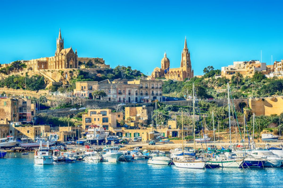 A sun-filled view of the Island of Gozo with yachts in the sitting in the water and beautiful buildings peering in the distance. 