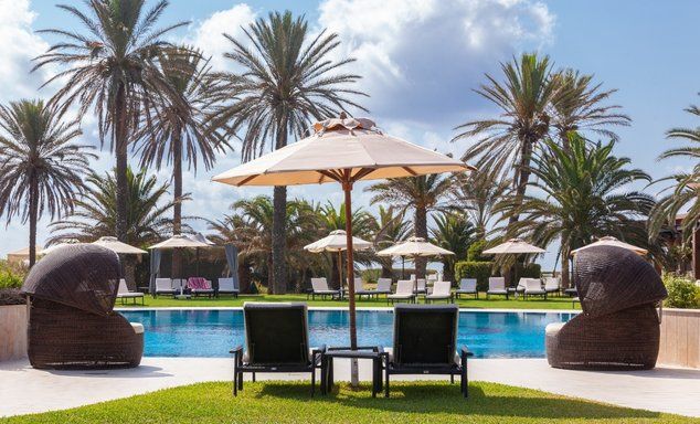 Two sun loungers and umbrella in front of an outdoor swimming pools with extra sun loungers placed on grass on the other side.
