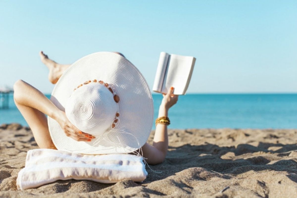 Woman with a big sun hat on reading a book on the beach.