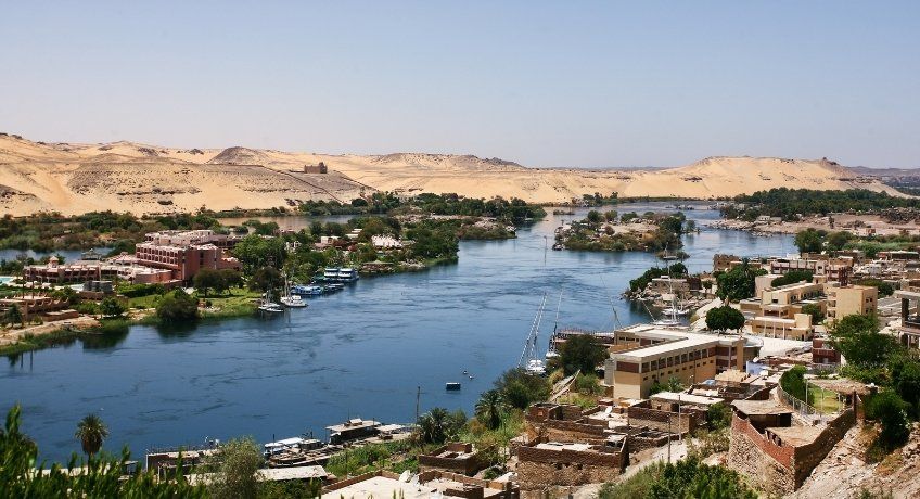 alt="Wide river valley: calm waterway through town, houses and boats along the banks, desert hills, clear blue sky."
