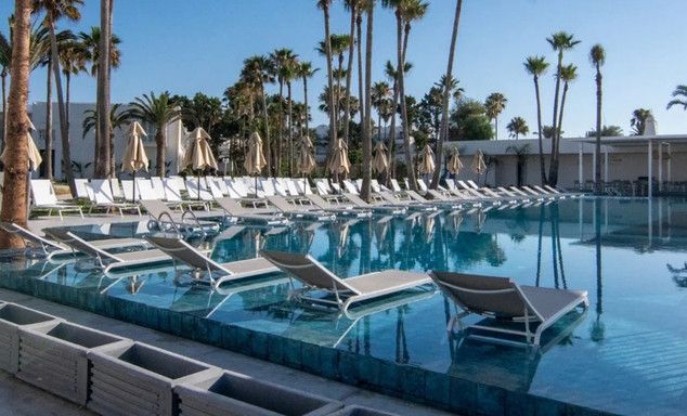 Beach resort pool with lounge chairs along the edge, palm trees, white cabanas, terrace, bright blue sky.