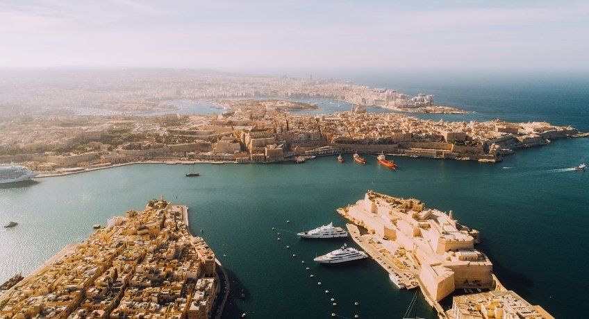 Aerial view of a coastal city: river and harbor, dense shoreline buildings, anchored boats, two curved breakwaters.