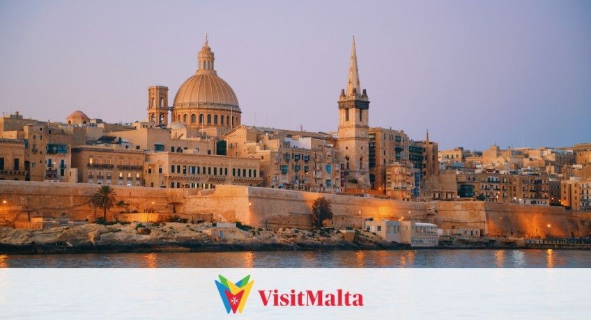 Sunset over Valletta's waterfront: St. Paul's Cathedral dome, a tall church tower, dense stone buildings, harbor.