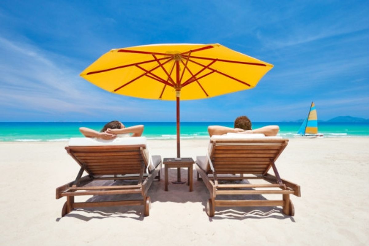 Two people sunbathing on sunloungers on the beach under an umbrella.
