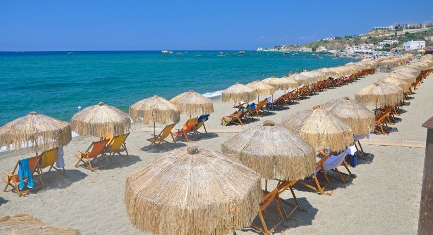 Sunny beach with rows of straw umbrellas and wooden loungers, turquoise water, coastal town on the right, clear sky.