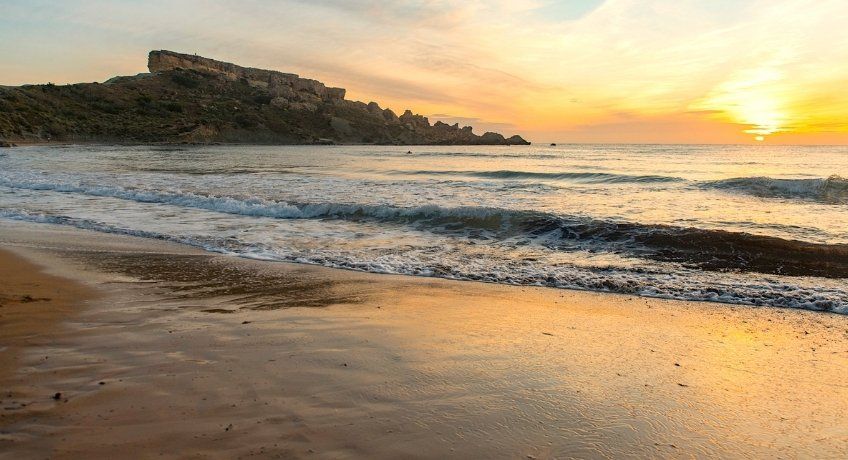 Golden-hour beach: gentle waves on wet sand, distant rocky headland, warm sunset sky with sun low over the sea.
