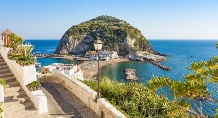 Coastal overlook with stone walkway and shrubs, small cove, turquoise water, rocky island with a building and pier.