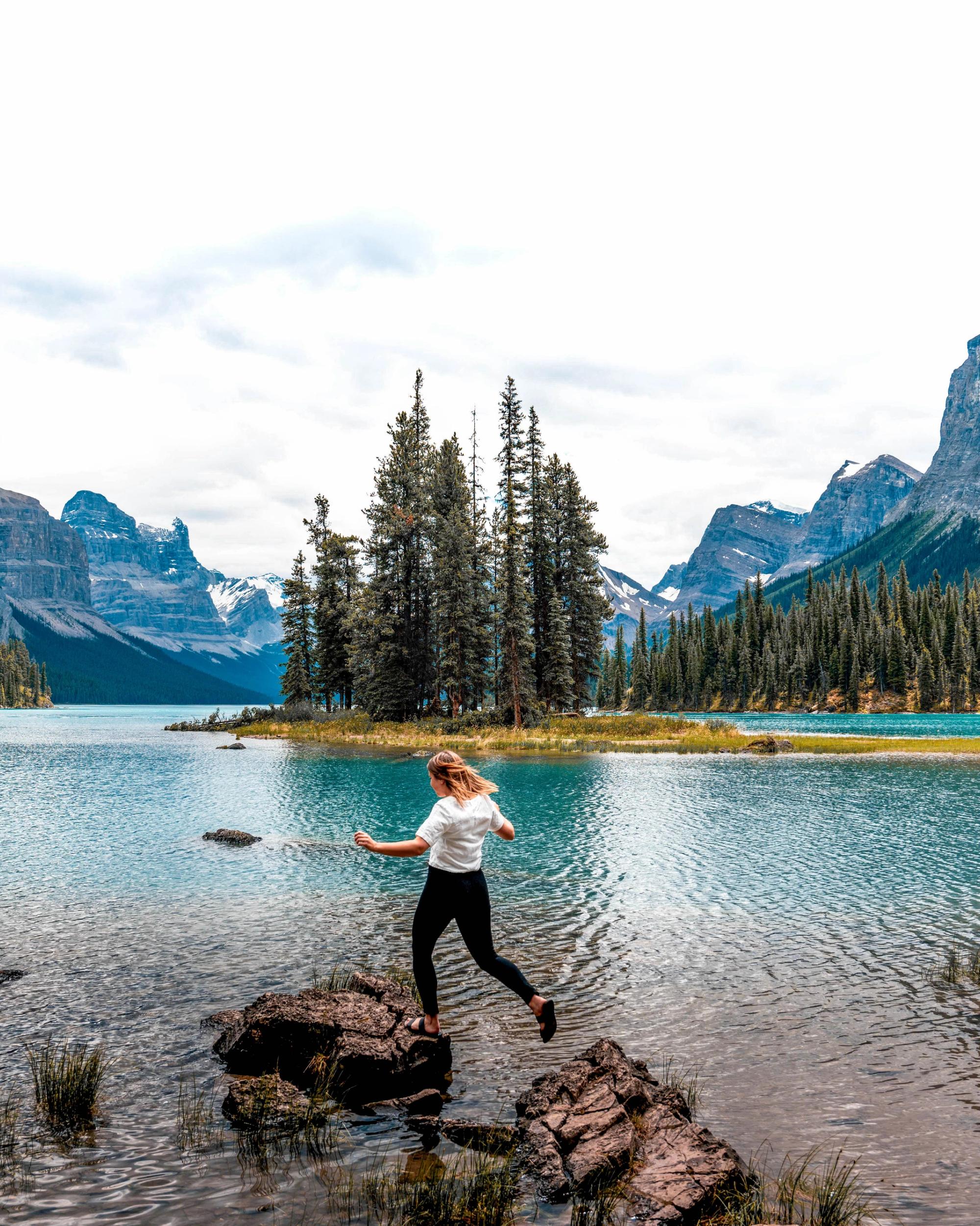 A hiker hopping across rocks in Maligne Lake, with Spirit Island behind