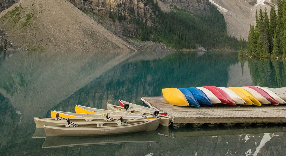 Canoes moored beside a dock of upturned coloured canoes on a calm mountain lake.