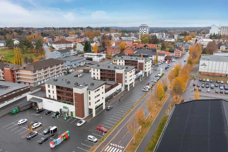Luftfoto av et byområde med moderne leilighetsbygg, parkeringsplasser, trerekke langs veien og omkringliggende småhusbebyggelse i høstfarger.