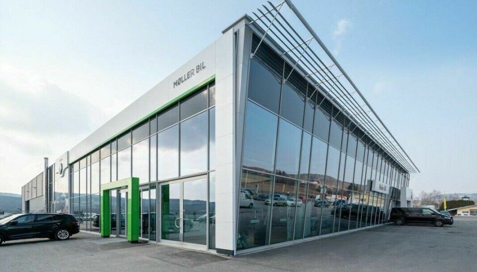 Moderne bilforhandlerbygning med stort glassfasadeparti, metallkonstruksjon og parkerte biler utenfor på et åpent uteområde.