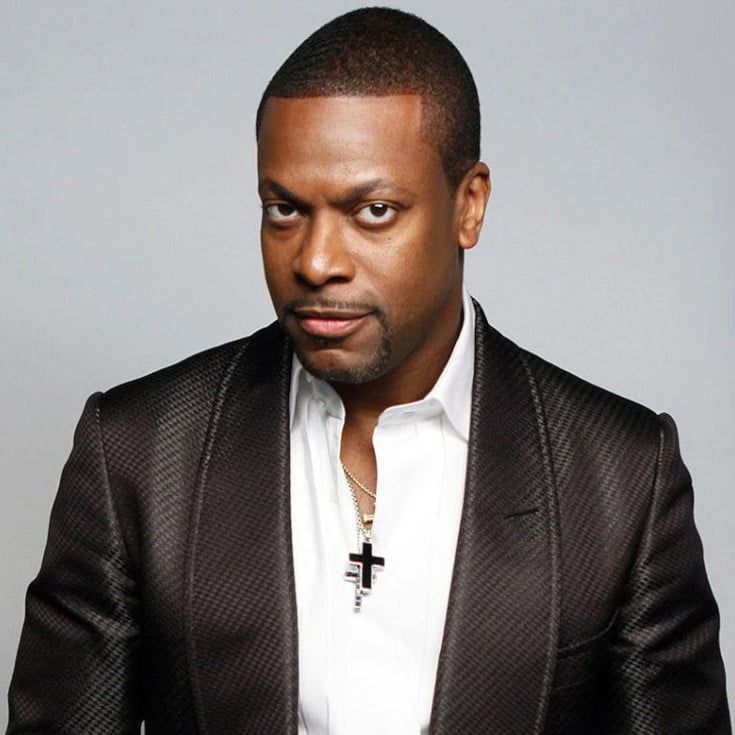 Chris Tucker wearing a crisp, white dress shirt with a deep, chocolate jacket, poses while standing for the camera. There is an added lavender frame around the photo.