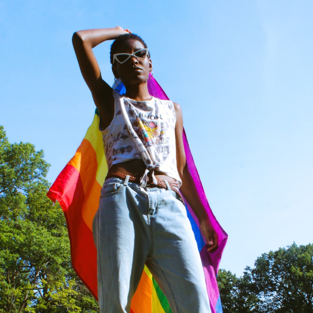 A young Black woman holds a rainbow flag behind her back. She's wearing sunglasses and looks cool in the summer sun.