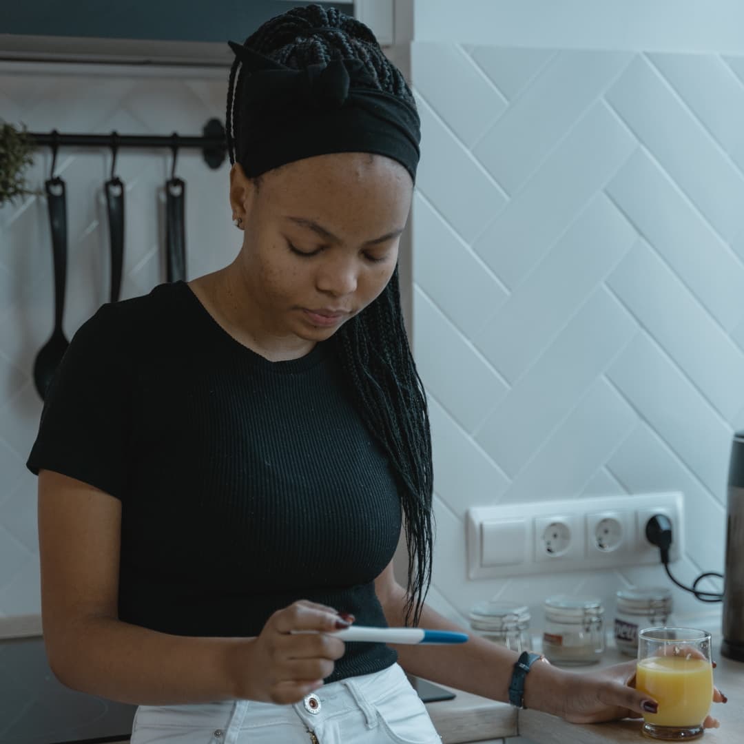 A Black woman reviewing an at-home pregnancy test. The test is in her left hand and a glass of orange juice is in her right hand.