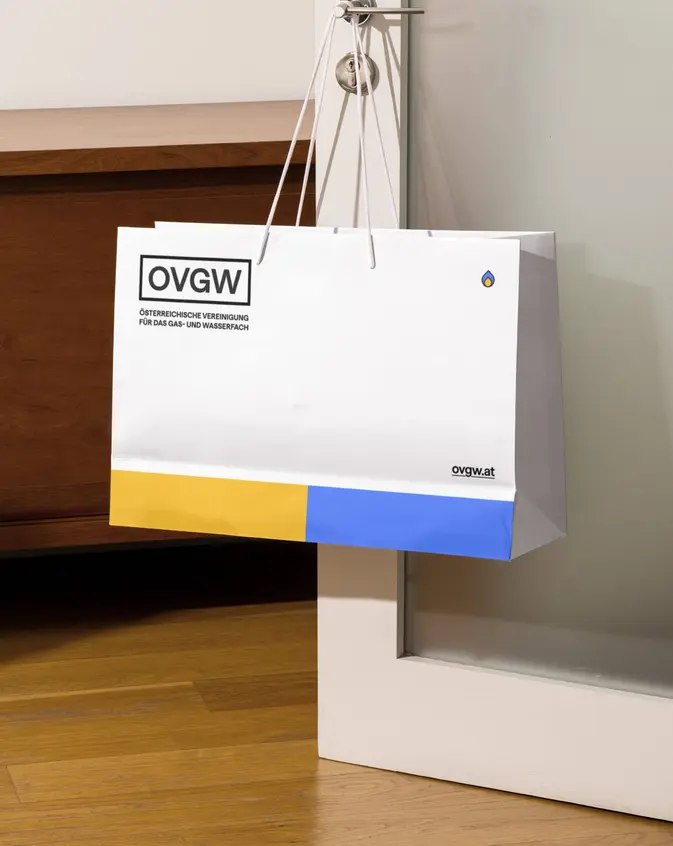 Eine weiße Papiertragetasche hängt an einer Türklinke und zeigt das OVGW-Logo