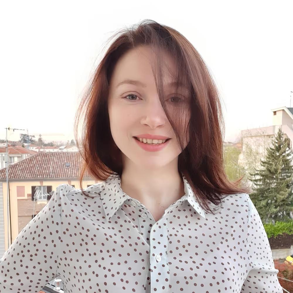 Smiling young woman with shoulder-length brown hair wearing a white shirt with brown polka dots, posing outdoors with buildings and trees in the background.