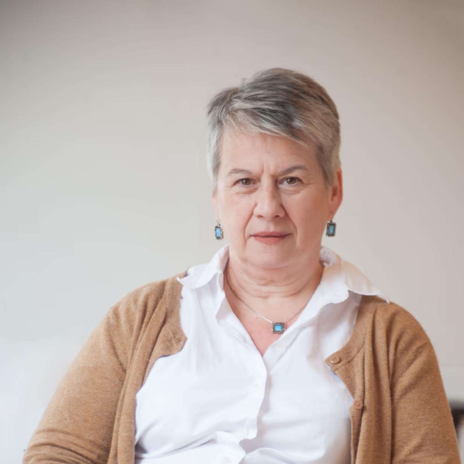 Middle-aged woman with short gray hair wearing a white shirt and brown cardigan with turquoise jewelry, sitting against a plain light background.