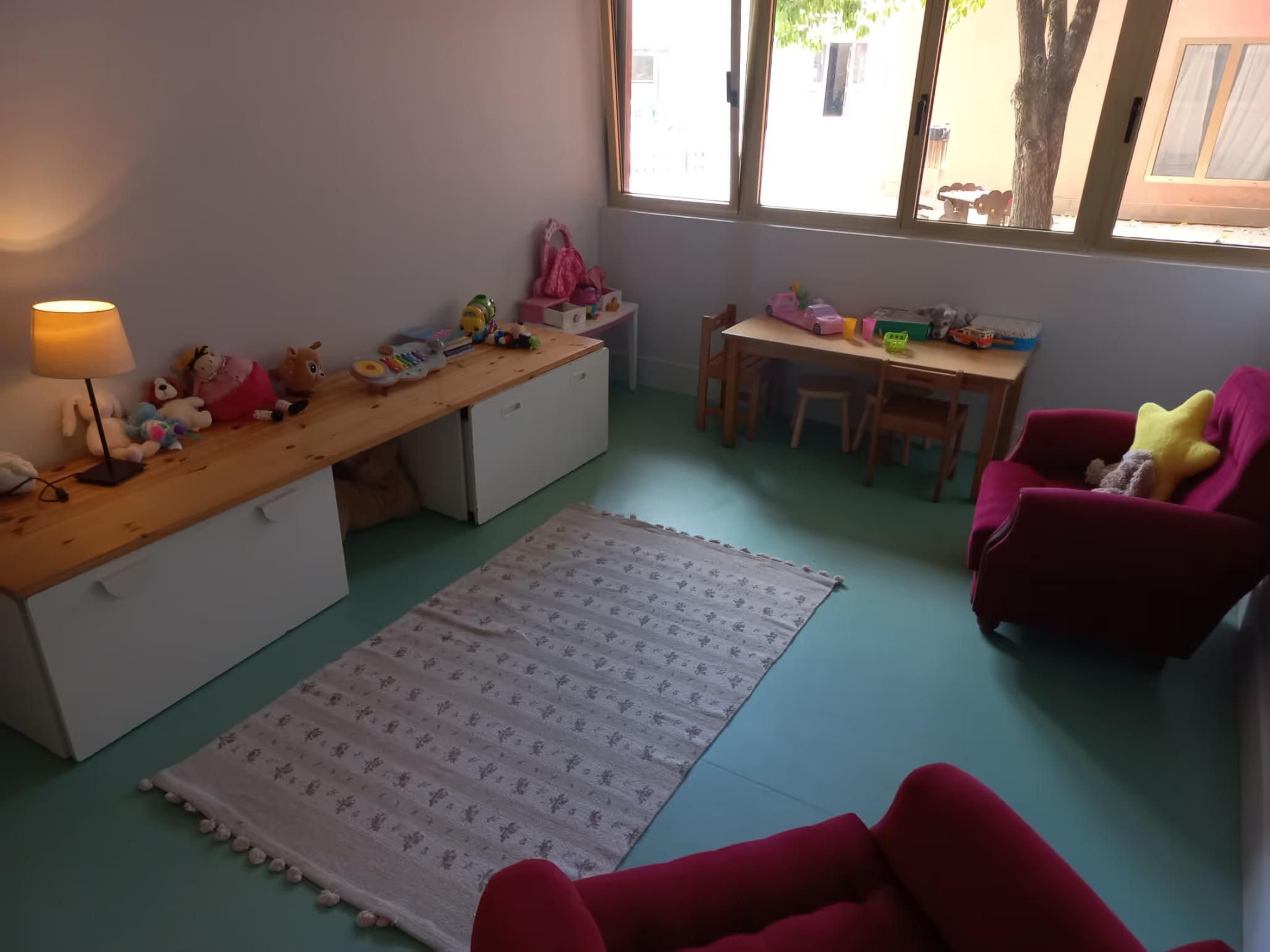 Children's playroom with toys on shelves, small table with chairs, and two red armchairs with cushions.