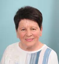 Portrait of a smiling elderly woman with short dark hair wearing a white top with blue and gray stripes on a light blue background.