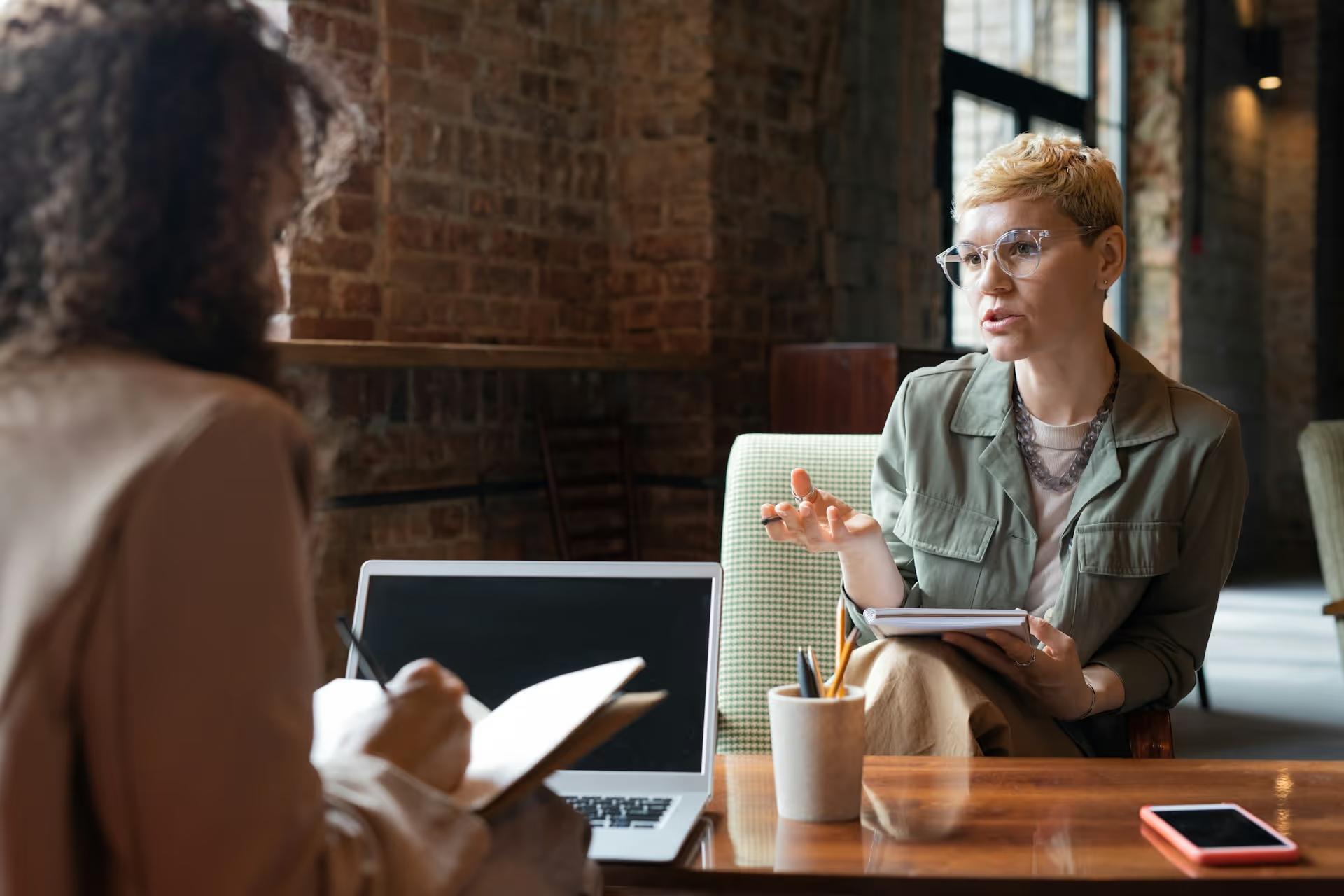 Two women talking across a café table with a laptop and notebook