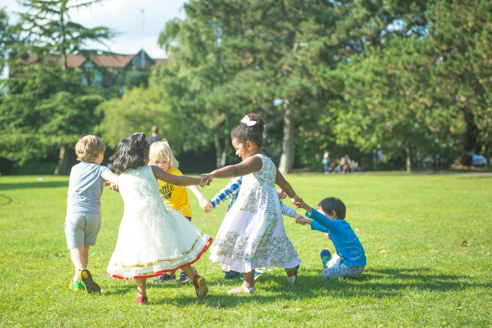 Young children hold hands in a circle on the grass in a park.