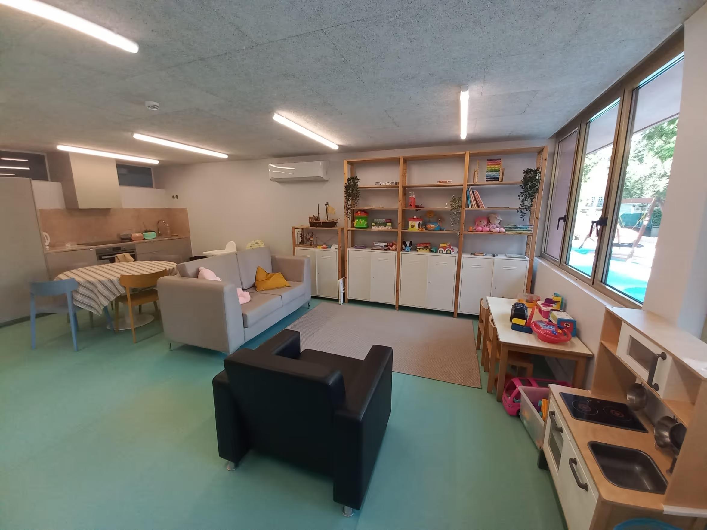 Bright playroom with toys, shelves, small tables, and seating near large windows and kitchenette.