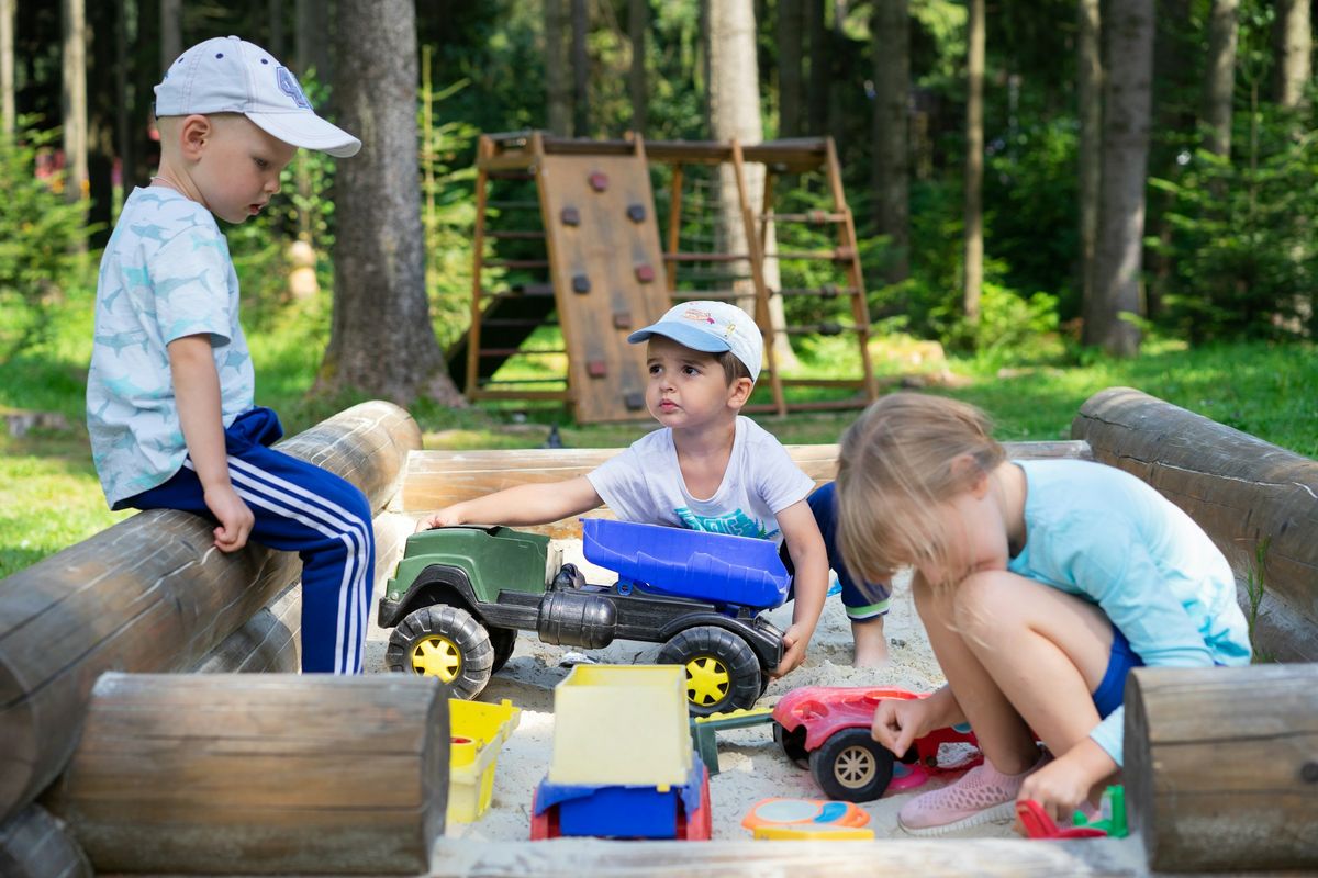 Three small children play with toy cars in an outdoor sand area.