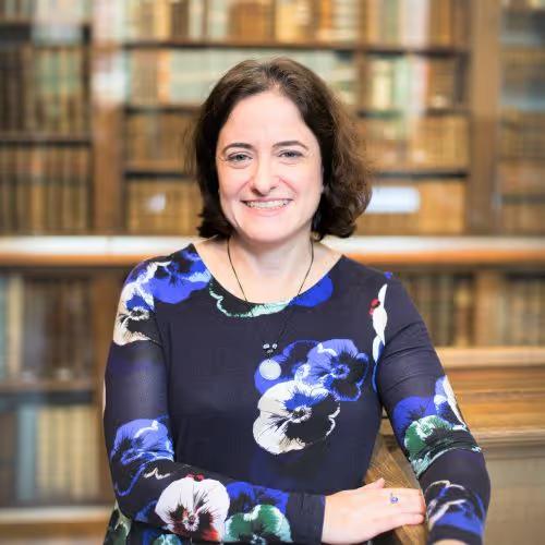Smiling woman with dark curly hair wearing a blue floral dress posed in front of a wooden bookshelf filled with books.