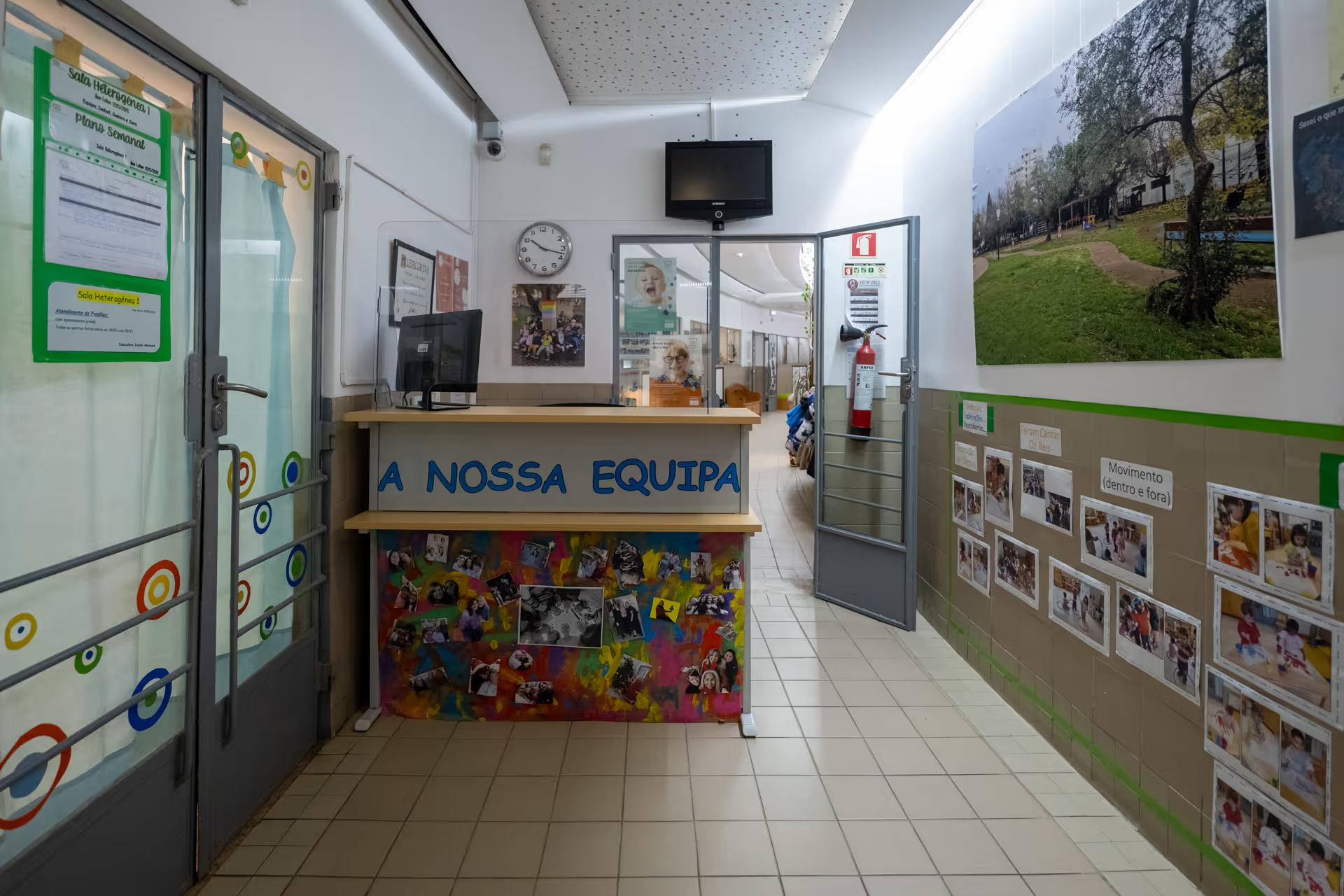 Reception area of a school with colorful desk labeled 'A NOSSA EQUIPA' and educational photos on walls.