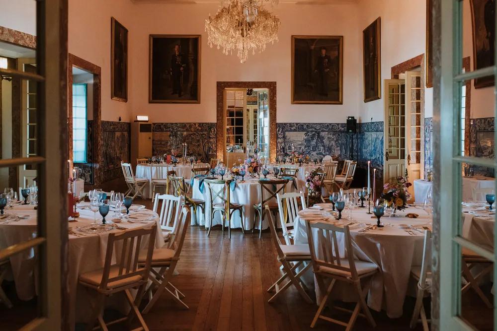 Elegant dining room with round tables, white chairs, floral centerpieces, and a crystal chandelier.