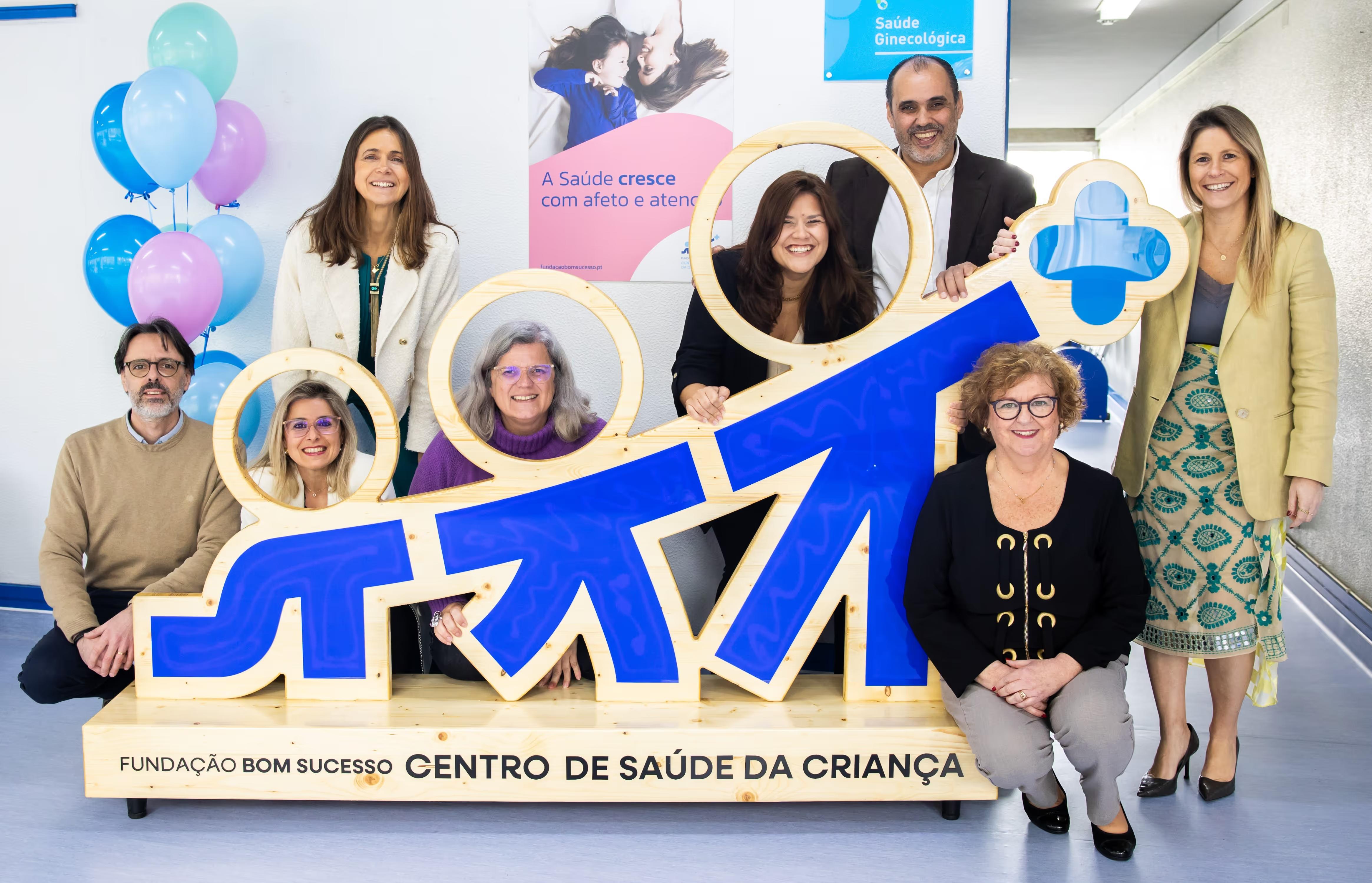 Eight smiling people posing around a large blue and wood Centro de Saúde da Criança sign with balloons nearby.