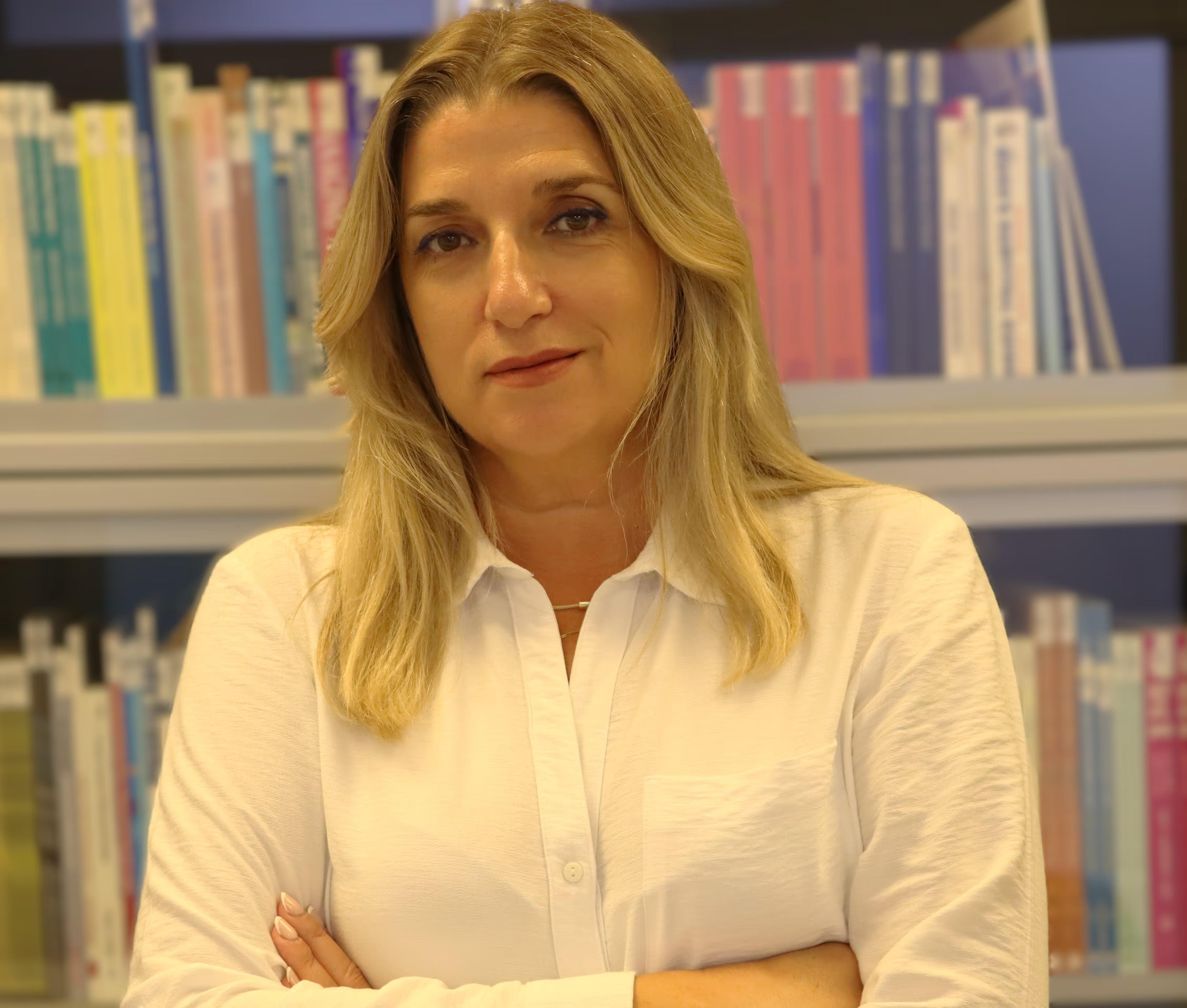 Middle-aged woman with blonde hair wearing a white shirt, standing with arms crossed in front of a bookshelf.