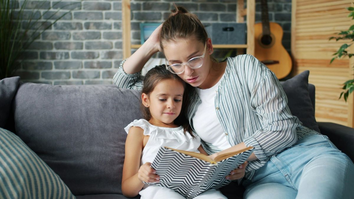 A mother and her young daughter read a picture book together on a sofa.