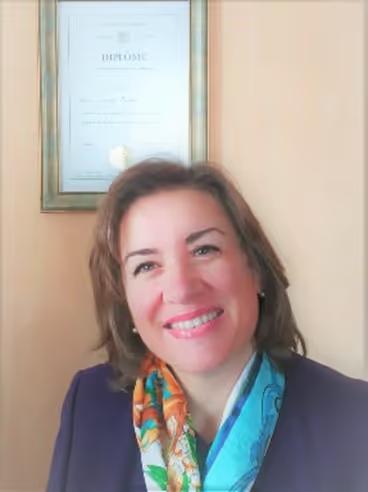 Smiling woman with shoulder-length brown hair wearing a colorful scarf and dark blazer, with a framed diploma on the beige wall behind her.