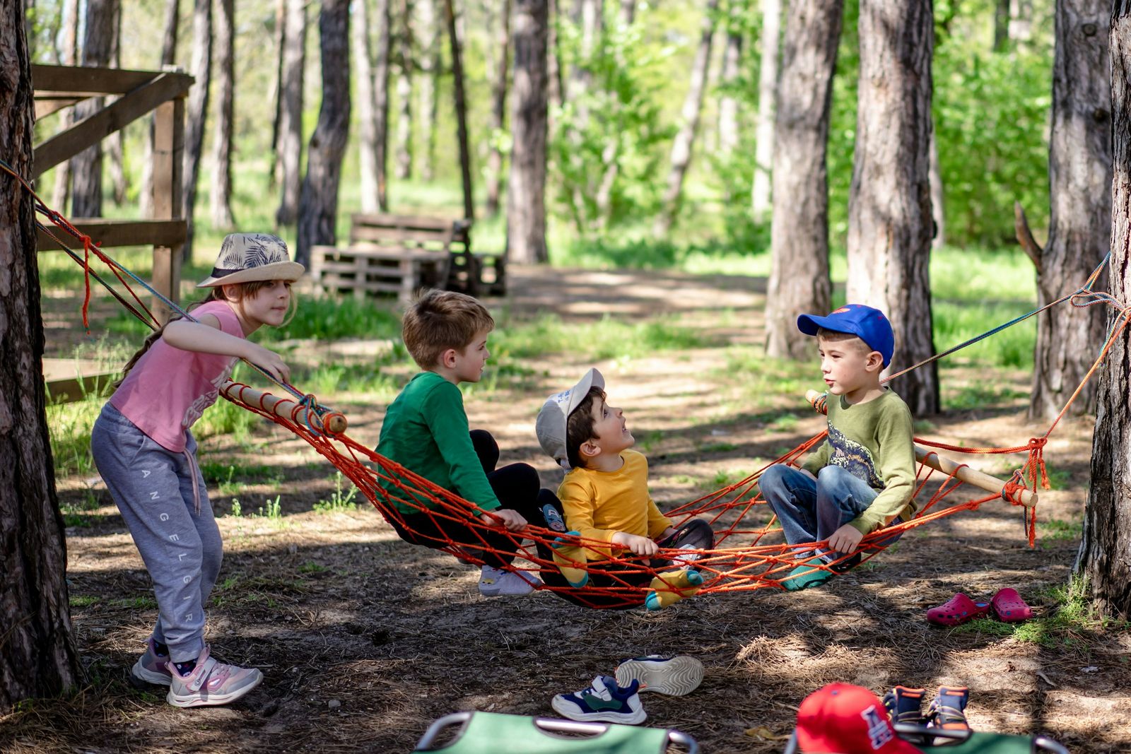 Young children play together around a hammock in a woodland playground.