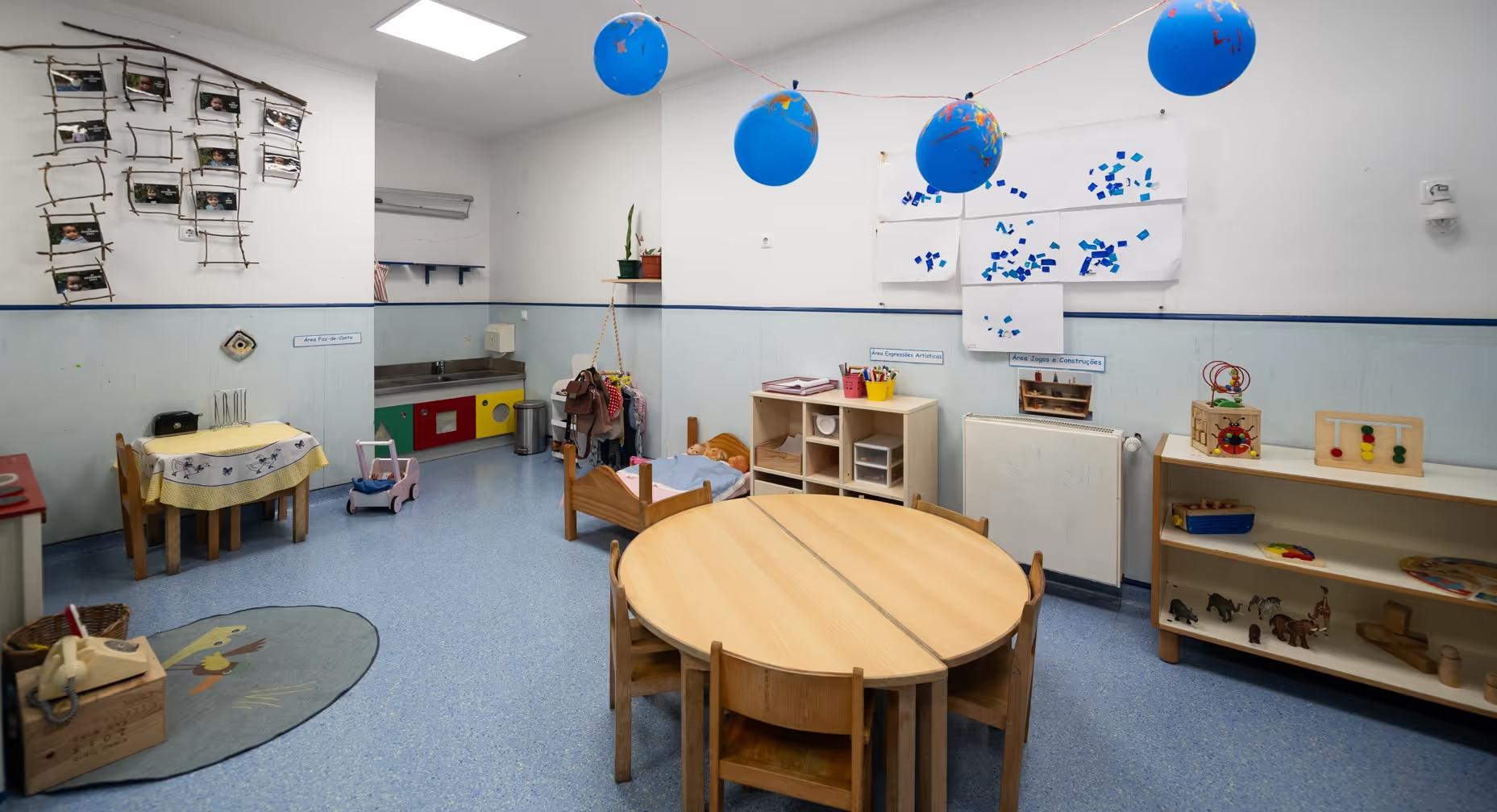 Empty preschool classroom with small tables, chairs, toys, blue floor, and balloons hanging from ceiling.