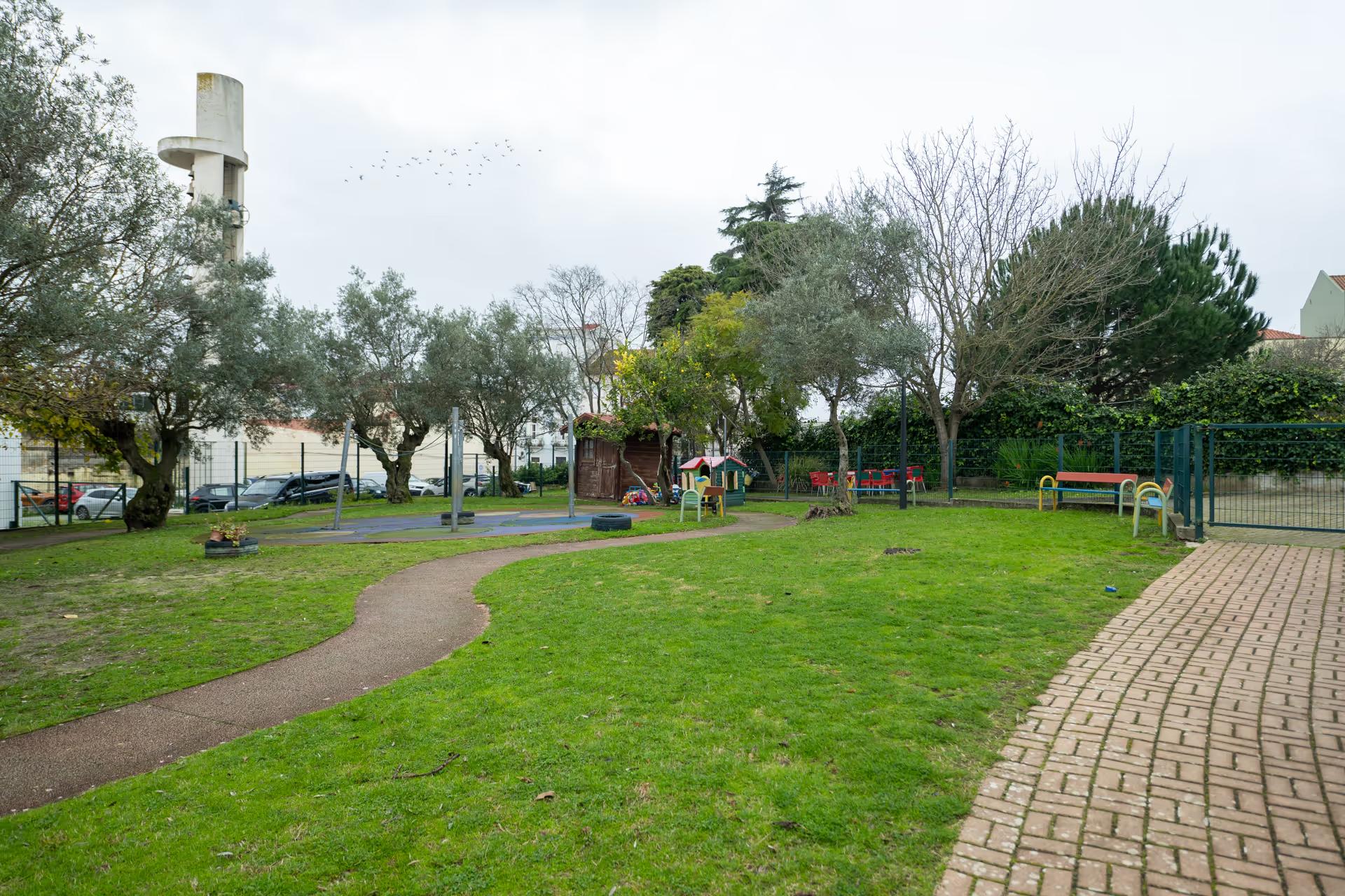 Park with green grass, playground equipment, benches, trees, and a paved walkway on an overcast day.