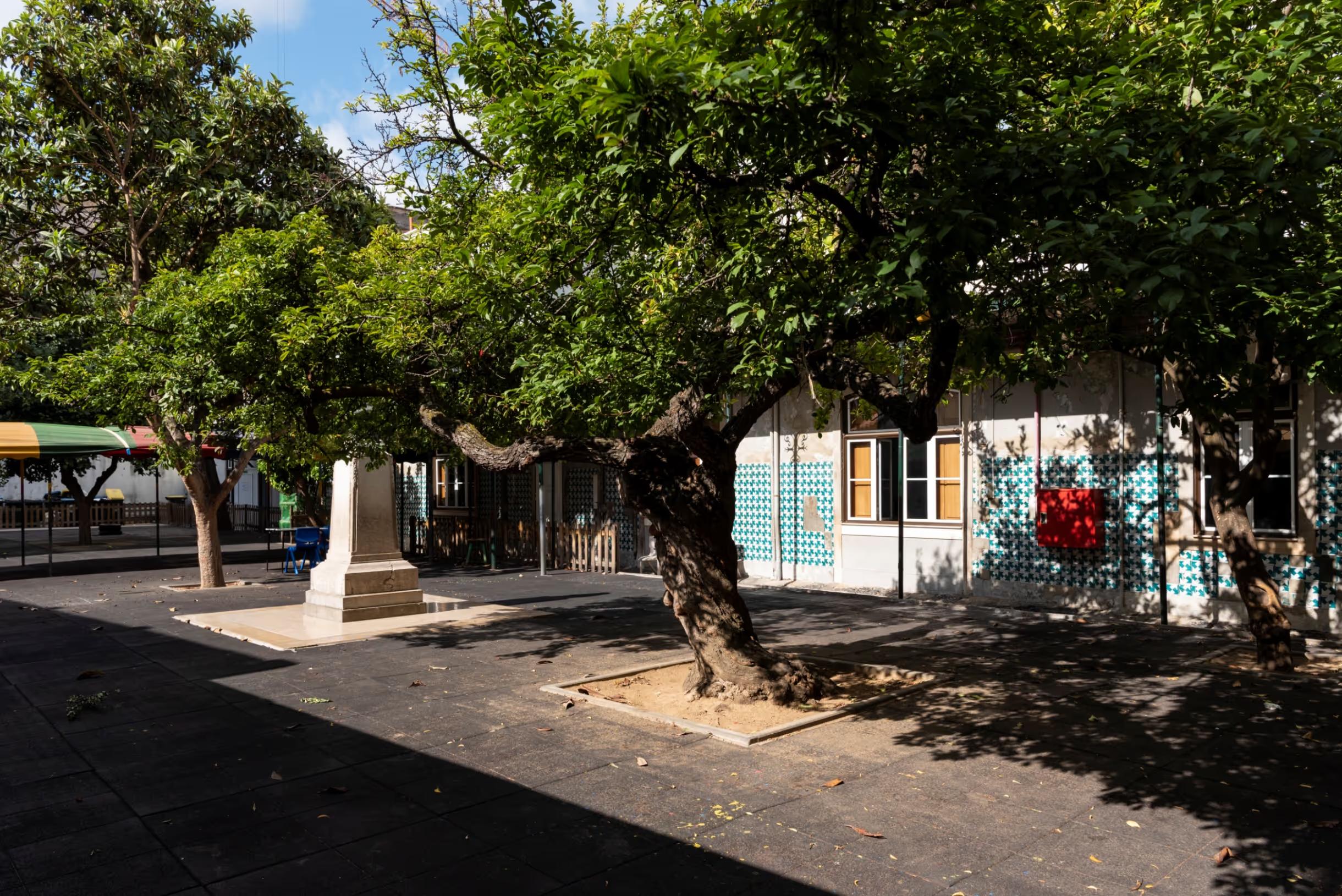 Shaded courtyard with trees, a stone monument, and a tiled building wall with windows.