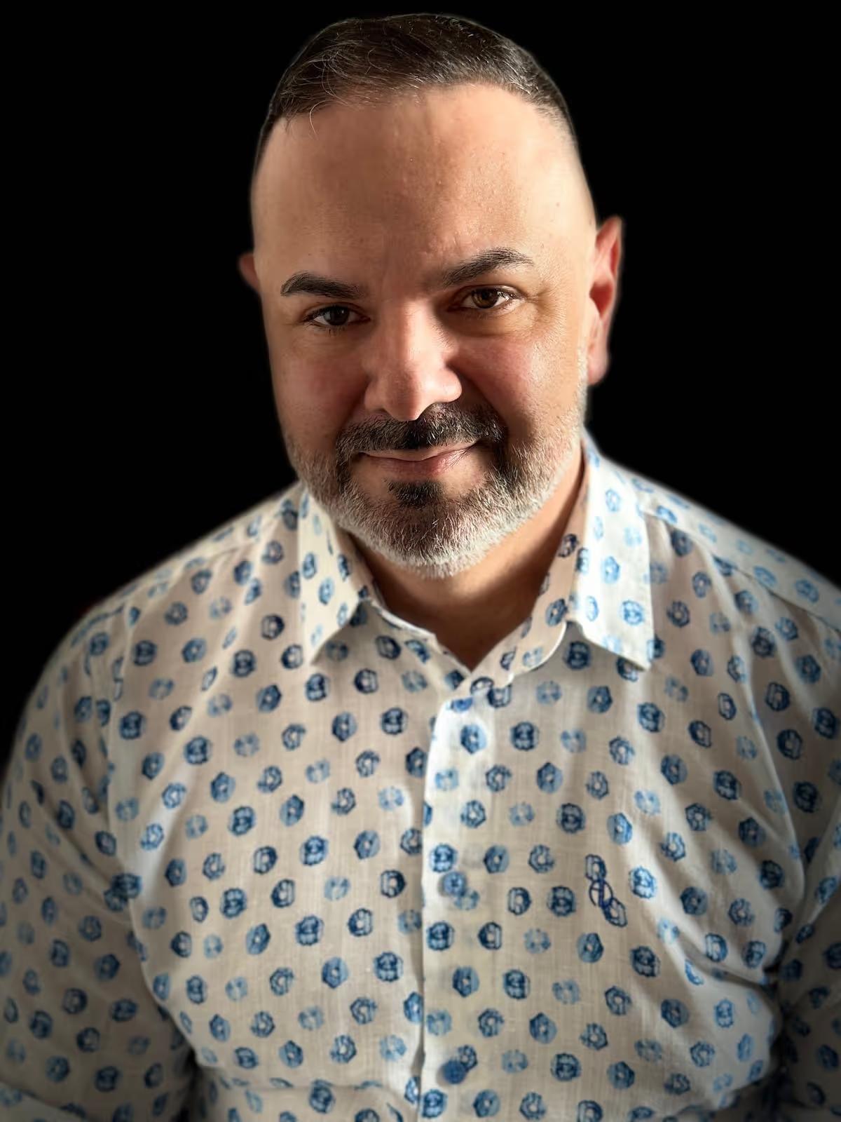 Man with short dark hair and beard wearing a white shirt with blue circular patterns against a black background.